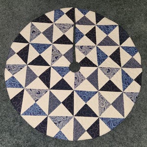 May include: Circular tree skirt with a geometric pattern. The skirt features a patchwork design of triangles in shades of blue, navy, and cream. The center has a hole for the tree trunk.