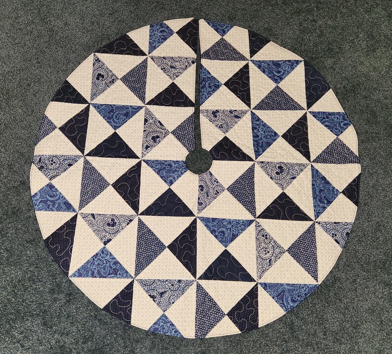 May include: Circular tree skirt with a geometric pattern. The skirt features a patchwork design of triangles in shades of blue, navy, and cream. The center has a hole for the tree trunk.