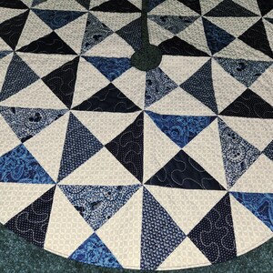 May include: Circular Christmas tree skirt with a geometric design. The skirt features triangles in various shades of blue and white, with patterned and solid fabrics. The design is quilted with white thread.