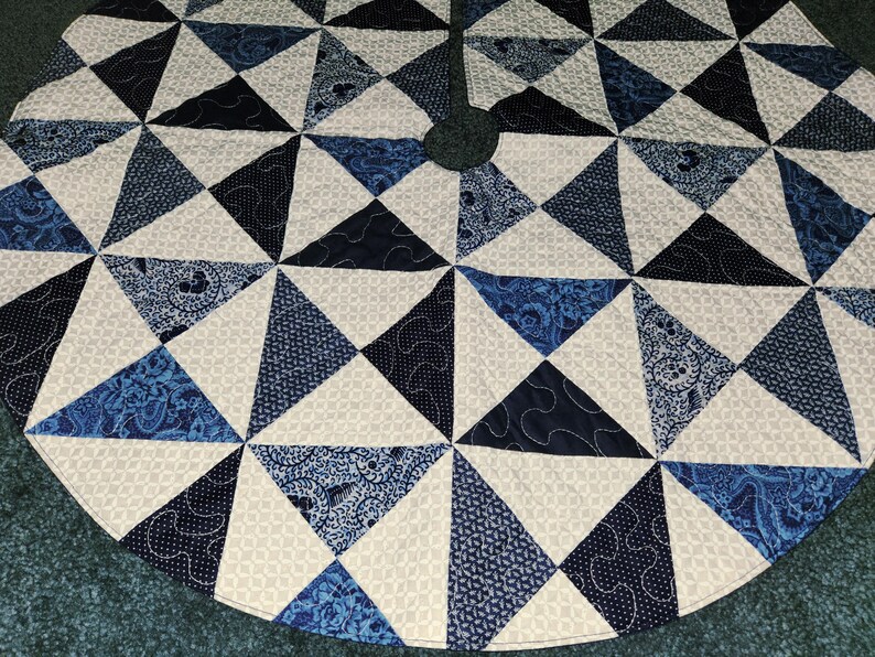 May include: Circular Christmas tree skirt with a geometric design. The skirt features triangles in various shades of blue and white, with patterned and solid fabrics. The design is quilted with white thread.