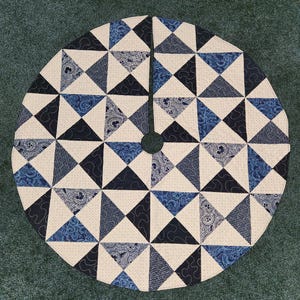 May include: Circular tree skirt with a patchwork design. The skirt features a repeating pattern of triangles in shades of blue and white. The center has a hole for the tree trunk.