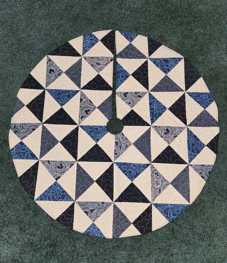 May include: Circular tree skirt with a patchwork design. The skirt features a repeating pattern of triangles in shades of blue and white. The center has a hole for the tree trunk.