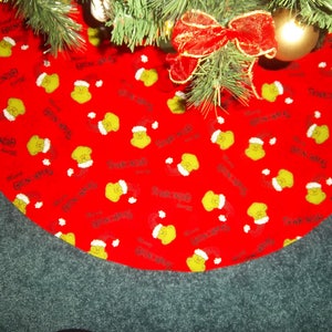 May include: Red Christmas tree skirt with a repeating pattern of a green character wearing a Santa hat and the words "Merry Grinchmas". The skirt is under a decorated Christmas tree with a red bow and gold ornaments.