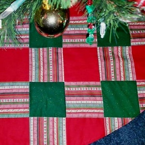 May include: A Christmas tree skirt with a patchwork design. The skirt features alternating red and green squares, with striped fabric accents in between. A gold ornament hangs from the tree.