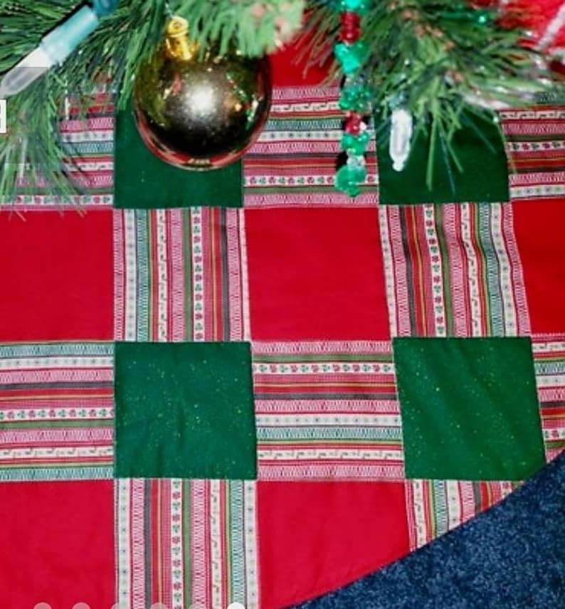 May include: A Christmas tree skirt with a patchwork design. The skirt features alternating red and green squares, with striped fabric accents in between. A gold ornament hangs from the tree.