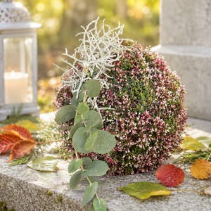 Könnte beinhalten: Ein kugelförmiges Blumenarrangement mit dichten rosa und grünen Heidekrautblüten, akzentuiert durch eine weiße zweigartige Struktur und einen herabhängenden Zweig mit runden, grünen Eukalyptusblättern für eine strukturierte Optik.