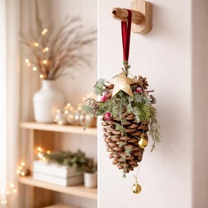 Op de afbeelding: Een decoratieve kerstversiering in de vorm van een grote dennenappel, versierd met een gouden ster, kunstgroen, rode bessen en kleine gouden belletjes. Het hangt aan een houten pin met een rood lint.