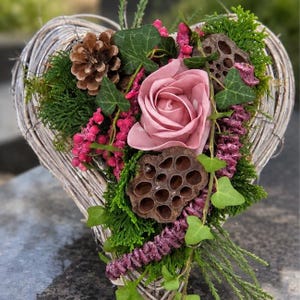 Könnte beinhalten: Ein herzförmiger Kranz aus geflochtenen, hellen Zweigen, verziert mit einer großen altrosa Rose, einem braunen Kiefernzapfen, einer getrockneten Lotus-Samenkapsel, grünem Efeu, leuchtend grünem Moos sowie kleinen rosa und violetten Blütenakzenten.