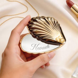 Op de afbeelding: Een gouden schelpvormige compacte spiegel met de naam "Nevaeh" gegraveerd op het spiegeloppervlak. De compact is tegen een achtergrond van satijnen stof gehouden, met een gouden ketting, parels, lippenstift en parfum in de buurt.