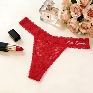 Peut inclure: Un string en dentelle rouge avec un petit nœud au centre et le texte brodé en blanc Mrs London sur le côté droit. Le vêtement est présenté à côté d'un flacon de parfum en verre, d'un tube de rouge à lèvres rouge et d'un bouquet de roses rose pâle.