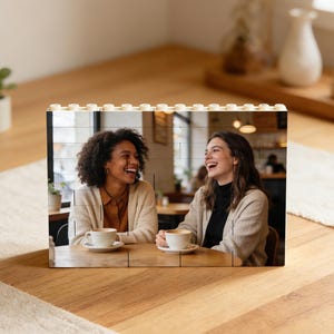 Puede incluir: Un marco de fotos de bloques de madera con una fotografía de dos mujeres riendo. El marco está construido con múltiples bloques, con una parte superior de color claro. Las mujeres están sentadas en una mesa con tazas de café.