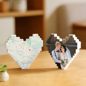 May include: Two heart-shaped decorative blocks. One features a map with a red heart, the other displays a wedding photo. Both are made of small, white blocks. The map block has text and a date. They sit on a wooden surface.