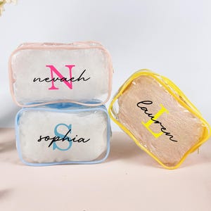 Puede incluir: Tres neceseres transparentes con nombres personalizados y letras iniciales. Una bolsa tiene un borde rosa y el nombre "Nevaeh" con una "N" rosa. Otra tiene un borde azul con "Sophia" y una "S" azul. La tercera tiene un borde amarillo con "Lauren" y una "L" amarilla.