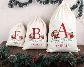 Puede incluir: Tres bolsas de regalo navideñas de color crema con letras rojas, ilustraciones de Papá Noel y las palabras "Merry Christmas". Las bolsas están personalizadas con los nombres: Emery, Brielle y Amelia. La decoración festiva rodea las bolsas.