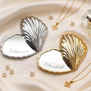 May include: A silver and gold heart-shaped compact mirror with a seashell design. The silver mirror is engraved with the name "Katherine", and the gold mirror is engraved with the name "Elizabeth". Gold necklaces and pearls are also in the image.