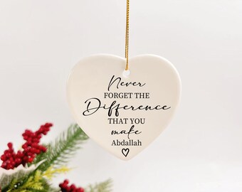 Pode incluir: Um enfeite de cerâmica em forma de coração, creme, com o texto "Never forget the difference that you make Abdallah" em escrita preta. Uma corda dourada é presa para pendurar. Bagas vermelhas e folhagem verde visíveis no canto inferior esquerdo.