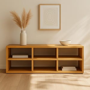 Peut inclure: Unité de rangement à six compartiments en bois clair, de conception minimaliste. Un bol blanc et une œuvre d'art abstraite encadrée ornent le dessus. Un vase avec de l'herbe de la pampa séchée et des piles de livres sont également visibles.