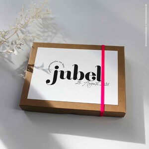 Könnte beinhalten: Eine braune Pappschachtel mit einer weißen Karte mit dem Wort "jubel" in schwarzer Schrift, akzentuiert durch ein leuchtend pinkfarbenes Band. Die Karte enthält auch das Datum "25. August 26".