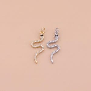 Peut inclure: Deux pendentifs en forme de serpent. L'un est doré, l'autre argenté. Les deux sont incrustés de petits cristaux transparents et ont des yeux noirs. Les pendentifs sont présentés sur un fond pêche clair.