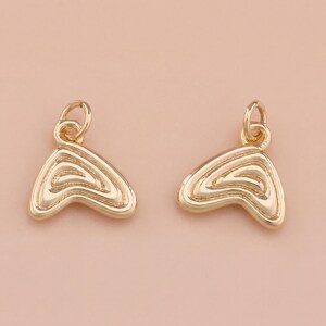 May include: Two gold-colored charms with a layered, abstract design. Each charm has a small loop at the top for attaching to a chain or other jewelry component. The charms are set against a soft, peach-colored background.