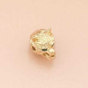 May include: A gold-coloured wolf head pendant with green gemstone eyes. The pendant features detailed fur texture against a soft pink background. Suitable for necklaces, bracelets, or other accessories. The pendant is approximately 2 cm in length.