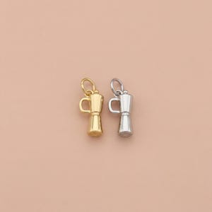 May include: Two small coffee pot charms, one gold-colored and one silver-colored, are displayed against a soft, peach-colored background. The charms are detailed with a handle and a spout, and are likely intended for jewelry making.