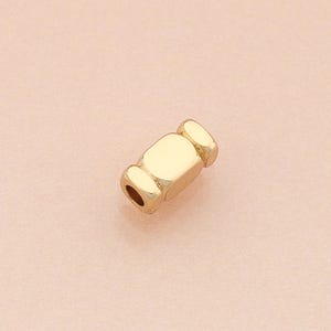 Puede incluir: Una cuenta dorada con un centro rectangular y extremos redondeados. La cuenta tiene un pequeño agujero que atraviesa el centro y probablemente se utiliza para la fabricación de joyas o manualidades. La cuenta se muestra sobre un fondo rosa claro.