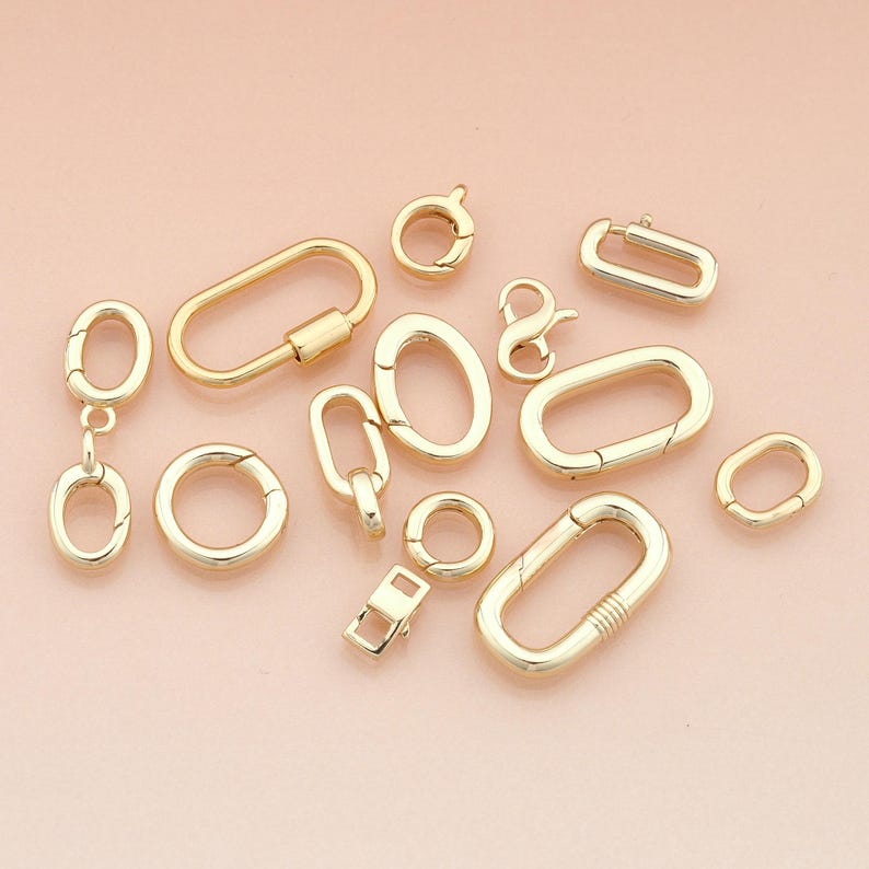 May include: A collection of gold-colored jewelry clasps and connectors in various shapes and sizes. The assortment includes oval, round, and rectangular designs, as well as a figure-eight clasp. The clasps are displayed on a light pink background.