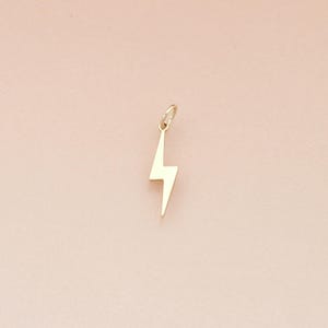 May include: A gold-colored lightning bolt charm with a small loop at the top. The charm is suspended against a soft pink background. The lightning bolt has sharp angles and a smooth surface, suggesting a pendant or jewelry piece.