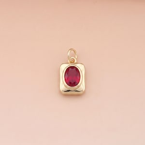 Könnte beinhalten: Goldfarbener Anhänger mit rechteckigem Rahmen und einem leuchtenden, oval geformten roten Edelstein in der Mitte. Der Anhänger hat oben eine kleine Schlaufe für eine Kette. Der Hintergrund ist ein sanftes Rosa.