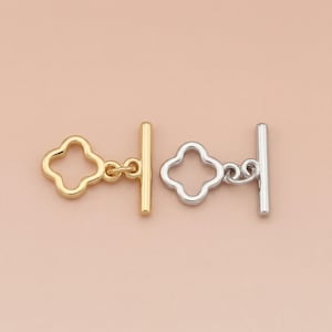 May include: Two metal toggle clasps, one gold-toned and one silver-toned, each featuring a clover-shaped loop and a straight bar. The clasps are designed for jewelry making and are displayed against a soft, neutral background.