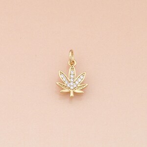 May include: A gold-colored pendant in the shape of a cannabis leaf. The leaf is detailed with clear, sparkling crystals. The pendant has a small loop at the top for attaching to a chain or bracelet. The background is a soft, peachy pink.