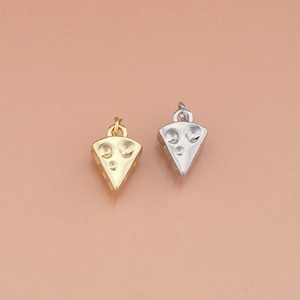 Puede incluir: Dos colgantes con forma de queso. Uno es de color dorado, el otro plateado. Ambos colgantes tienen tres agujeros circulares y un pequeño lazo en la parte superior para sujetarlos a una cadena o pulsera. El fondo es de color melocotón claro.