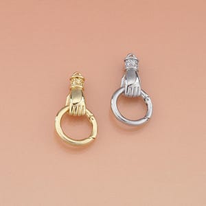 May include: Two metal clasp charms, one gold-toned and one silver-toned, each featuring a hand holding a circular ring. The hand has intricate detailing and a decorative top. The clasps are likely for jewelry or accessories.