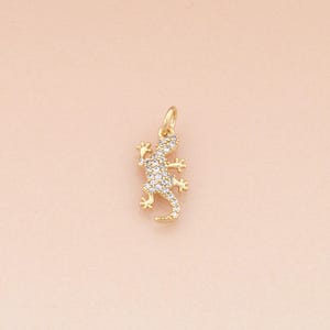 Op de afbeelding: Een goudkleurige hagedis hanger met heldere, sprankelende kristallen. De hagedis is gedetailleerd met kleine pootjes, een gekrulde staart en een lus voor een ketting. De hanger staat tegen een zachtroze achtergrond.