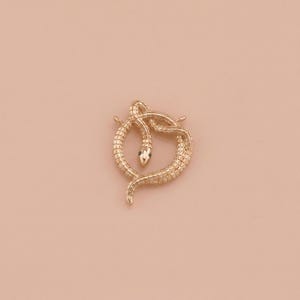 Pode incluir: Broche dourado em forma de cobra com um olho de gema verde. A cobra está enrolada num design circular. O broche está sobre um fundo de cor pêssego claro.