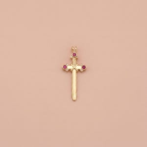 Op de afbeelding: Een goudkleurige hanger in de vorm van een zwaard. De hanger heeft drie rode edelstenen en een gedraaid ontwerp op het handvat. De zwaardhanger staat op een effen, lichtroze achtergrond.