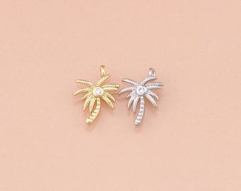 May include: Two palm tree pendants, one gold-toned and one silver-toned, each featuring a small, clear gemstone at the center. The pendants are detailed with intricate leaf designs and a loop for attaching to a chain or bracelet. The background is a soft, peachy-pink.