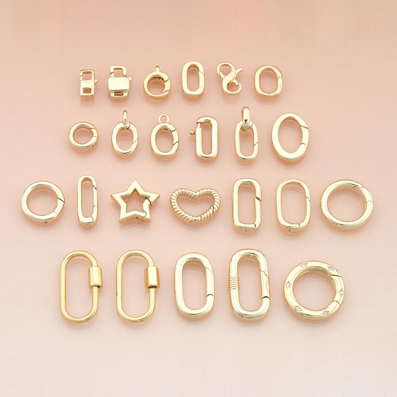 May include: A collection of gold-colored jewelry clasps and connectors in various shapes, including circles, ovals, rectangles, and a star and heart design. The clasps are displayed on a light pink surface.