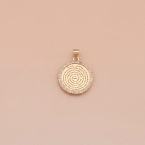 May include: Gold-coloured pendant with a circular design. The pendant has a ring of small, clear stones around the edge. The centre of the pendant has a spiral pattern with text. A small loop is at the top for a chain.
