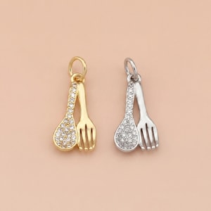 May include: Two pendant charms, one gold-toned and one silver-toned, each featuring a spoon and fork design. The spoon is encrusted with small, clear gemstones. The fork has three tines. The background is a solid, light peach color.