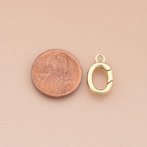 May include: A gold-colored oval clasp with a small loop at the top, next to a penny. The clasp is open, revealing the interior. The penny shows the profile of a man's face and the words "In God We Trust."