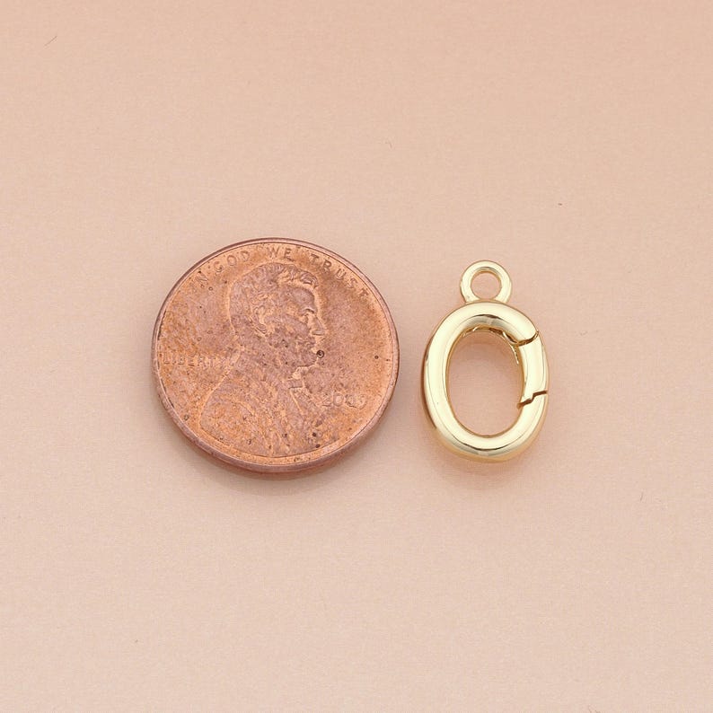 May include: A gold-colored oval clasp with a small loop at the top, next to a penny. The clasp is open, revealing the interior. The penny shows the profile of a man's face and the words "In God We Trust."