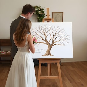 K&ouml;nnte beinhalten: Eine Alternative zum Hochzeitsg&auml;stebuch mit einem handgemalten Baumdesign auf einer wei&szlig;en Leinwand. Der Baum hat braune &Auml;ste und einen Stamm. Die Namen "Iryna & Patrick" und das Datum "05.05.2025" sind darunter geschrieben. Die Leinwand wird auf einer Holzstaffelei ausgestellt.