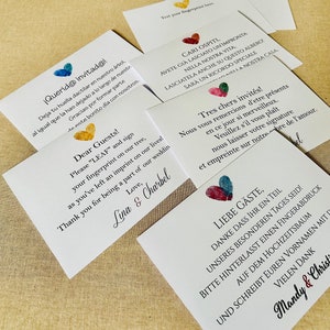 K&ouml;nnte beinhalten: Wei&szlig;e Karten mit bunten herzf&ouml;rmigen Fingerabdruck-Designs und Text in mehreren Sprachen. Die Karten laden die G&auml;ste ein, ihre Fingerabdr&uuml;cke auf einem Baum zu hinterlassen. F&uuml;r eine Hochzeit.