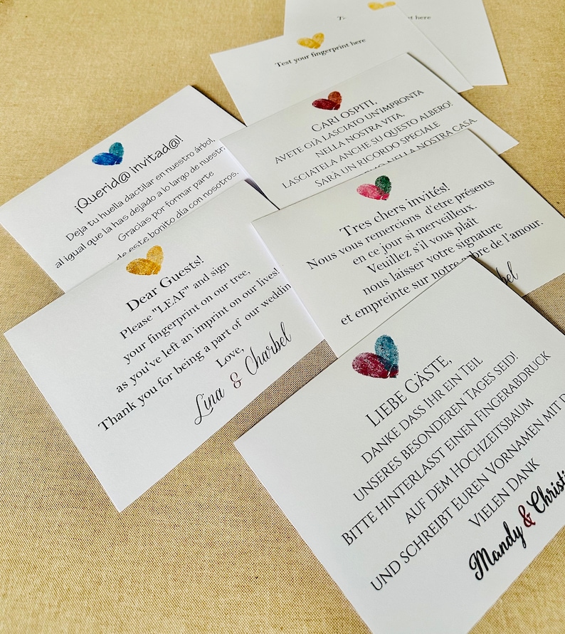 K&ouml;nnte beinhalten: Wei&szlig;e Karten mit bunten herzf&ouml;rmigen Fingerabdruck-Designs und Text in mehreren Sprachen. Die Karten laden die G&auml;ste ein, ihre Fingerabdr&uuml;cke auf einem Baum zu hinterlassen. F&uuml;r eine Hochzeit.