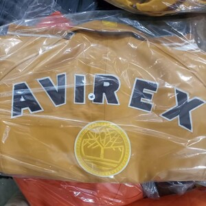 May include: Mustard-colored jacket with the word "AVIREX" in large, black, block letters across the back. A circular patch with a tree design is below the text. The jacket is in a clear plastic bag.