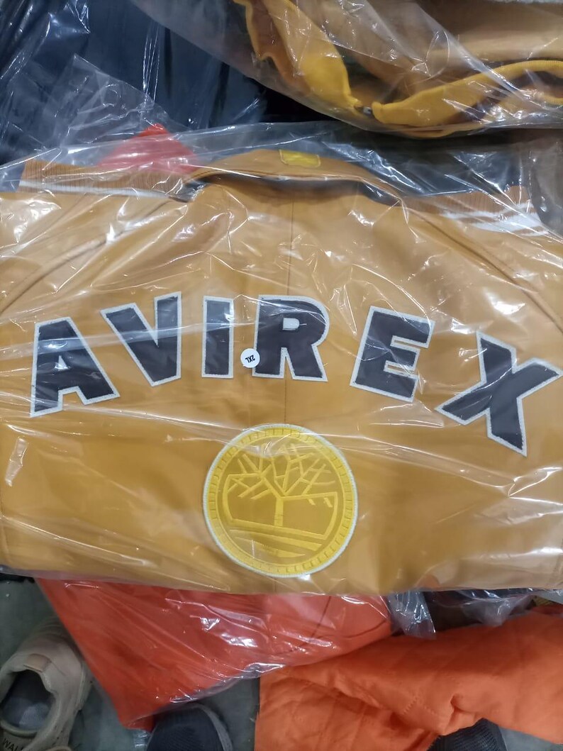 May include: Mustard-colored jacket with the word "AVIREX" in large, black, block letters across the back. A circular patch with a tree design is below the text. The jacket is in a clear plastic bag.