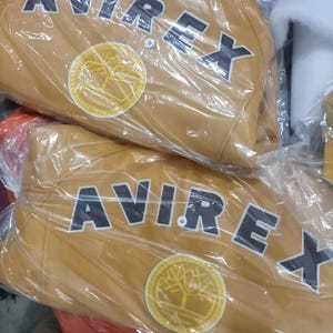 May include: Two tan leather jackets are visible, each wrapped in clear plastic. The jackets feature the word "AVIREX" in large, black block letters across the back, above a circular emblem with a tree design.