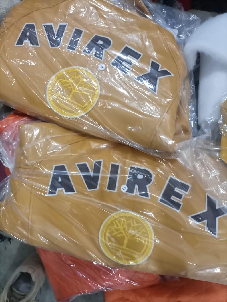 May include: Two tan leather jackets are visible, each wrapped in clear plastic. The jackets feature the word "AVIREX" in large, black block letters across the back, above a circular emblem with a tree design.